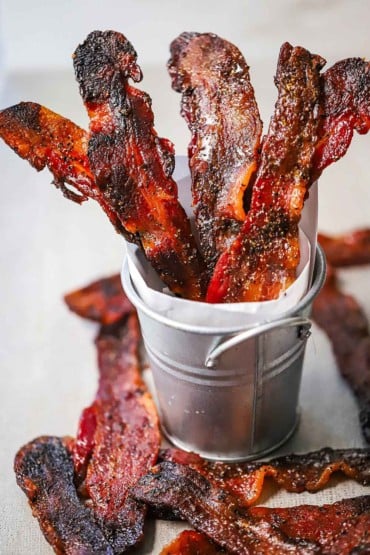 Five strips of maple pepper bacon sitting upright in a small silver pale with extra strips of bacon laying around the base of the pale.