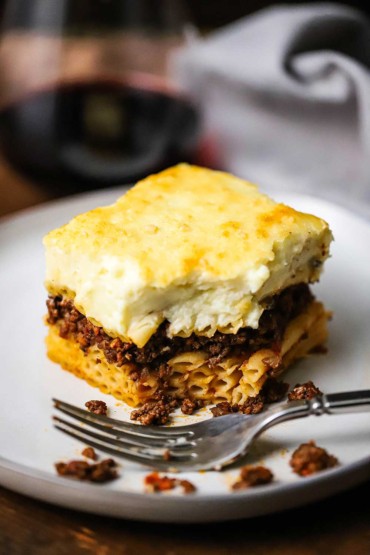 A straight-on view of a slice of Greek pastitsio with a bite take out of it on a white dinner plate with a fork nearby.