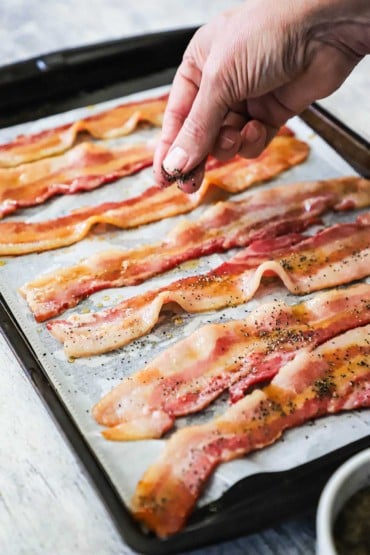 A person sprinkling coarsely ground black pepper over the tops of strips of bacon sitting on parchment paper on a baking pan.