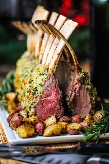 A straight-on view of two roast rack of lamb persillade that are on a platter with the bones interlocking above the chops forming a cathedral-looking roast.