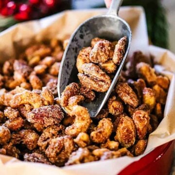A close-up view of a person holding a metal scooper filled with spiced nuts over a Christmas tin filled with more of the nuts.