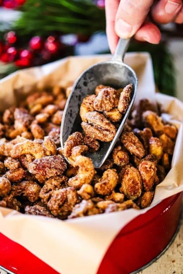A close-up view of a person holding a metal scooper filled with spiced nuts over a Christmas tin filled with more of the nuts.