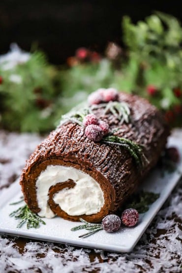 A straight-on view of a yule log that is sitting on a rectangular white platter and is topped with sugared cranberries and sprigs of rosemary all topped with sprinkled powdered sugar.