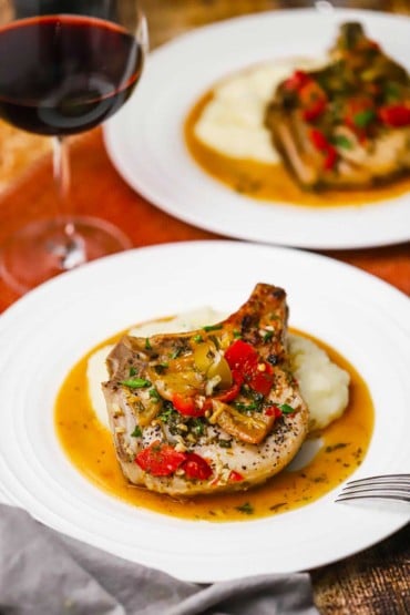 A straight-on view of two white dinner plates filled with Italian-style smothered pork chops both resting in a light brown sauce and a glass of red wine nearby.