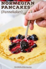 A person using a small spoon to spread blueberry jam over a thin Norwegian pancake (pannekaker) on a white plate.