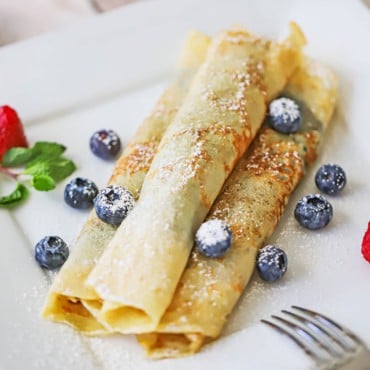 An overhead view of three Norwegian pancakes that have been rolled up and stacked on a plate with fresh blueberries scattered around and dusted with powdered sugar.