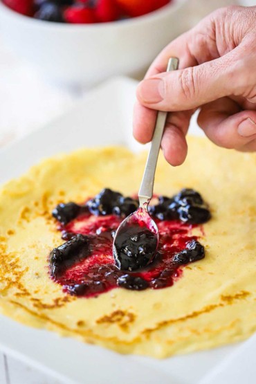 A person using a small spoon to spread blueberry jam over a thin Norwegian pancake (pannekaker) on a white plate.