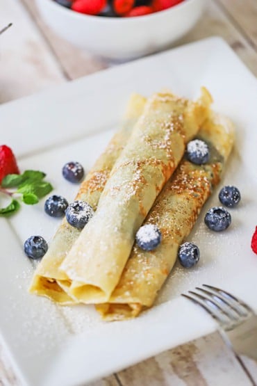 An overhead view of three Norwegian pancakes that have been rolled up and stacked on a plate with fresh blueberries scattered around and dusted with powdered sugar.
