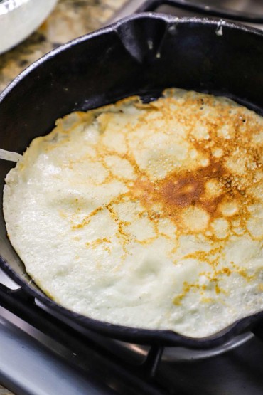 An overhead view of a thin crepe-like Norwegian pancake that is lightly browned and being cooked in cast-iron skillet.