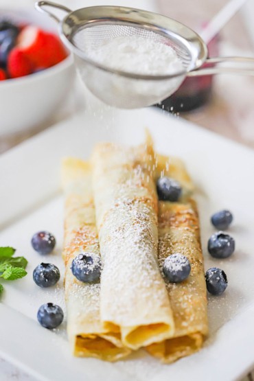 A small sieve being used to sprinkle powdered sugar over the tops of three rolled Norwegian pancakes with blueberries scattered around.