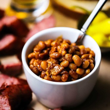 A straight-on view of slow cooker baked beans in a small individual white bowl surrounded by slices of cooked sausage and a glass of beer nearby.