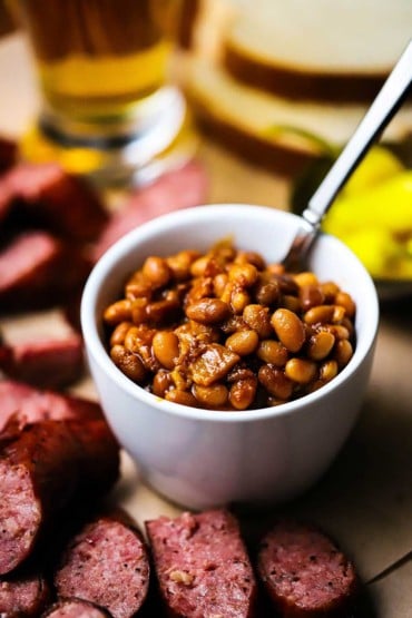 A straight-on view of slow cooker baked beans in a small individual white bowl surrounded by slices of cooked sausage and a glass of beer nearby.