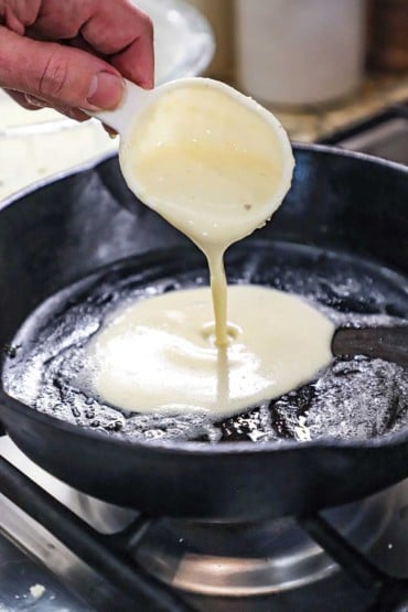 A person pouring Norwegian pancake batter from a plastic measuring cup into a black cast-iron skillet that has a thin layer of melted butter on the bottom.