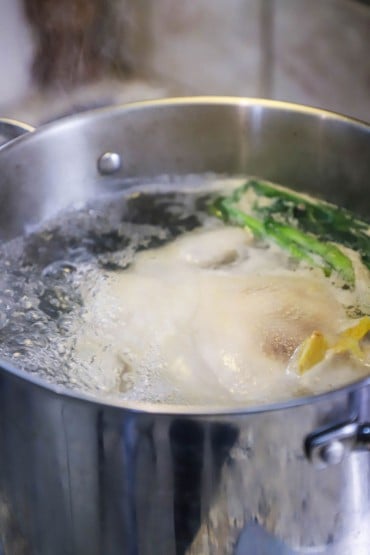 A close-up view of a whole chicken in a large stock pot of boiling water with scallions and fresh ginger simmering, too.