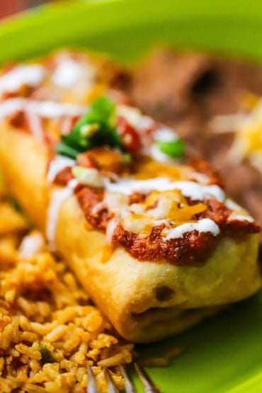 A close-up view of a deep-fried beef chimichanga with a layer of ranchero sauce and melted Colby jack cheese on top.