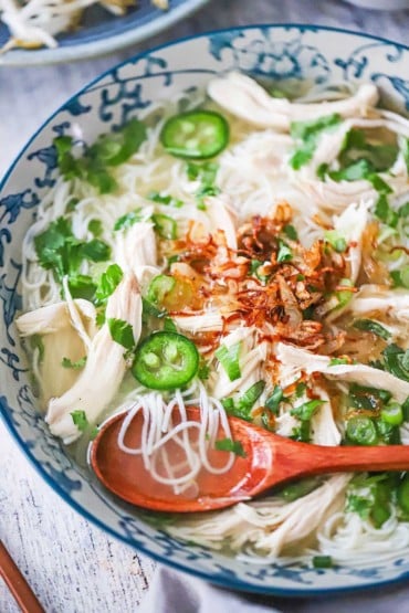 An overhead view of a colorful bowl filled with chicken pho that is garnished with sliced jalapeños, cilantro, and fried shallots, with a wooden spoon inserted.