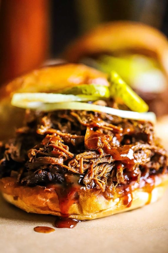 A close-up view of a slow cooker chopped brisket sandwich with barbecue sauce drizzled over the top of the meat with a couple of pickle slices and a thinly slice of onion on top, too.