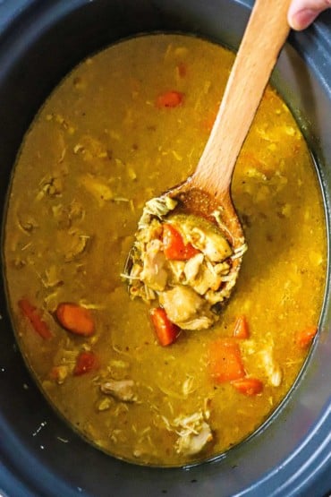 An overhead view of a 6-quart slow-cooker filled with a coconut cream sauce with cooked chicken, carrots, and sweet potatoes, with a large wooden spoon holding up chicken and sauce.