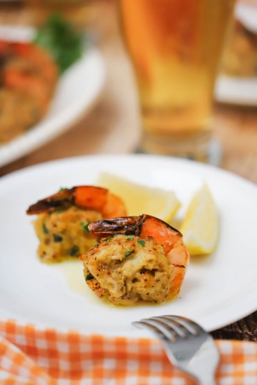 Two crab-stuffed shrimp sitting on a small white plate with a tall glass of beer in the background.