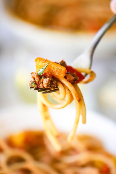 A close-up view of a fork that is being held up and is holding bucatini pasta and ground turkey with yellow sweet peppers.