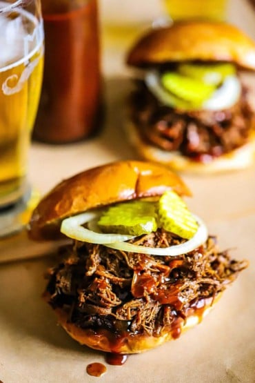 An overhead view of two slow cooker chopped brisket sandwiches that are topped with pickles and sliced onion and a glass of beer nearby.