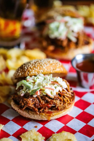 A straight-on view of a slow cooker pulled pork sandwich sitting on a red checkered sandwich wrapper with kettle chips scattered around it.