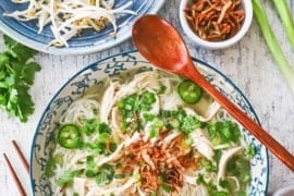 An overhead view of a bowl of chicken pho garnished with cilantro, peppers, and fried shallots, sitting next a platter of additional garnishes, including lime wedges and mung bean sprouts.
