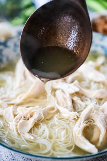 A large wooden ladle being used to pour chicken broth into a bowl of shredded cooked chicken and vermicelli noodles.