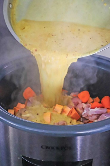 A coconut curry sauce being poured from a large saucepan into a Crock-Pot filled with chunks of uncooked chicken and cut-up carrots and sweet potato.