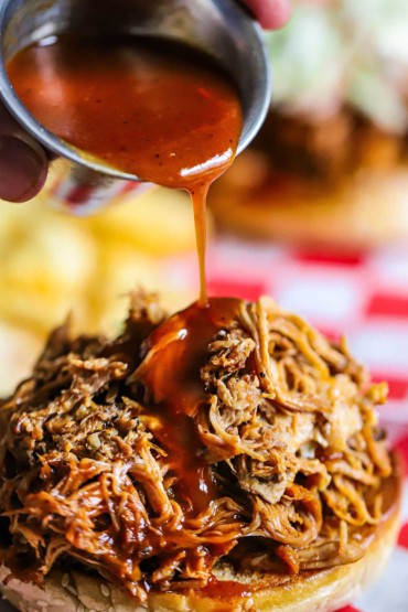 A person drizzling Carolina barbecue sauce over a pile of slow cooker pulled pork sitting on the bottom portion of a hamburger bun.