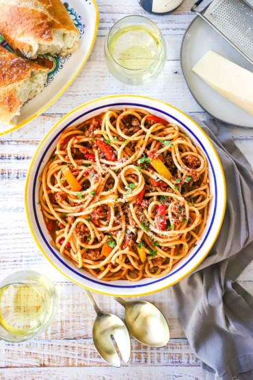 An overhead view of a large pasta bowl filled with pasta with turkey and sweet pepper ragu and surrounded by a glass of white wine and Italian bread.