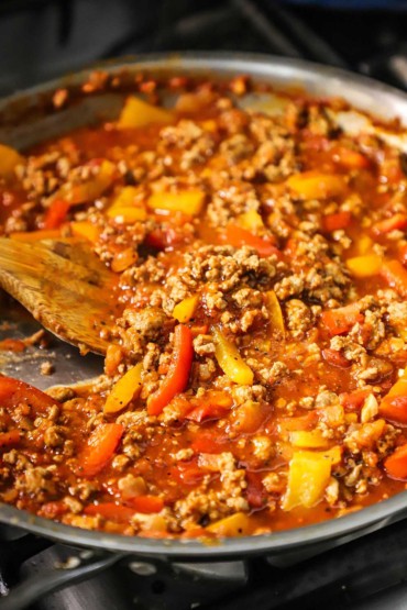 A close-up view of a large silver skillet filled with a ground turkey and sweet pepper ragu with a wooden spatula inserted in the middle of the sauce.