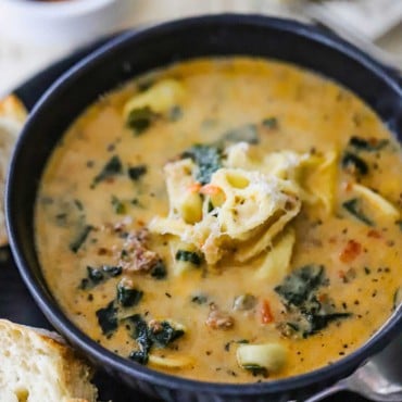 A close-up view of a blue soup bowl filled with creamy tortellini soup topped with grated parmesan cheese with two chunks of Italian bread sitting nearby.