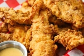 A straight-on view of a basket that is lined with a red-checkered piece of wax paper and is filled with a pile of crispy chicken tenders and a small container of honey mustard dipping sauce.