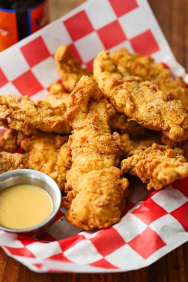 A straight-on view of a basket that is lined with a red-checkered piece of wax paper and is filled with a pile of crispy chicken tenders and a small container of honey mustard dipping sauce.