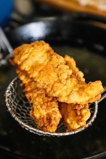 A straight-on view of three chicken tenders that are sitting in a metal spider over a skillet filled with hot oil.