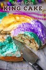 A straight-on view of a colorful Mardi Gras King cake that has a slice missing where the cinnamon filling is visible and a large knife is next to the cake.