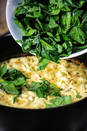 Chopped fresh spinach being dumped from a white bowl into a cast-iron pot filled with simmering creamy tortellini soup.