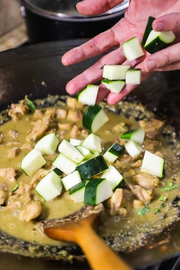 A person dropping bite-sized pieces of zucchin into a wok filled with a simmering Thai green curry chicken sauce.