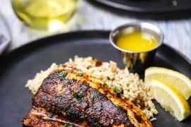 A straight-on view of a blackened redfish filet resting against a bed rice pilaf with two lemon wedges and a small vessel of melted butter nearby.