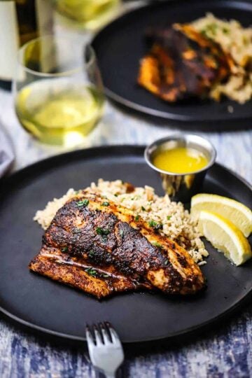 A straight-on view of a blackened redfish filet resting against a bed rice pilaf with two lemon wedges and a small vessel of melted butter nearby.