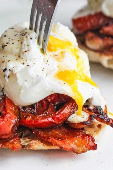 A fork being used to puncture a poached egg sitting on top of roasted tomato slices, bacon, and a toasted English muffin with the egg yolk streaming from the egg.