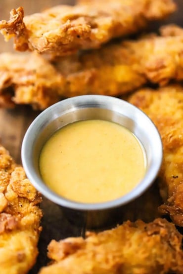 A close-up view of a small silver condiment vessel filled with homemade honey mustard dipping sauce surround by chicken tender.
