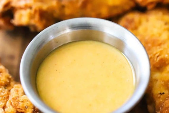 A close-up view of a small silver condiment vessel filled with homemade honey mustard dipping sauce surround by chicken tender.