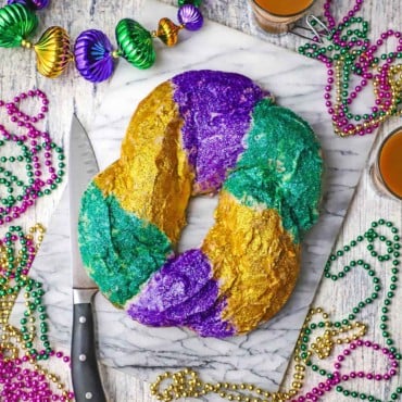 An overhead view of a circular Mardi Gras King Cake that is covered with shiny bright purple, green, and gold edible glitter and is surround by Mardi Gras beads.