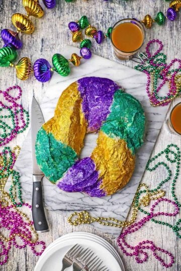 An overhead view of a circular Mardi Gras King Cake that is covered with shiny bright purple, green, and gold edible glitter and is surround by Mardi Gras beads.