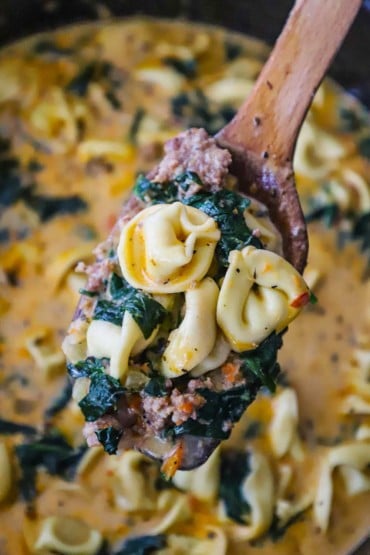 A close-up view of a wooden spoon holding a large serving of tortellini soup with wilted spinach and chunks of cooked ground sausage.