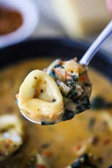 A close-up view of a cooked tortellini sitting and chunks of cooked spinach on a silver spoon over a pot of soup.