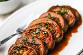 A straight-on view of a white oval platter filled with slices of stuffed flank steak that has been covered in a wine reduction sauce.