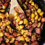 A close-up overhead view of a cast-iron skillet filled with corned beef hash with a wooden spoon inserted in the middle of it all.